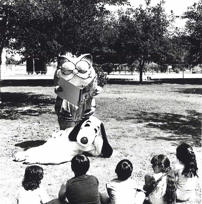 Black and white image of the cartoon characters Snoppy & Garfield as mascots reading to children sitting outside at a park