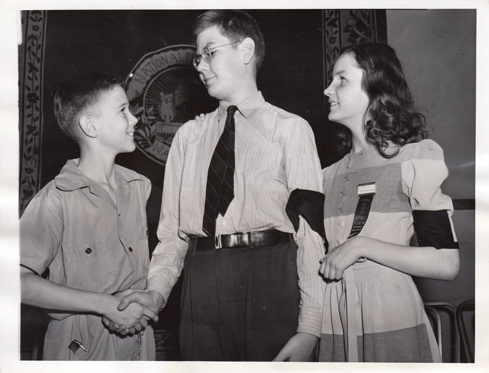 Black & White Picture Of The 1941 Spelling Bee Champion Shaking a Boys Hand While A Woman Watches
