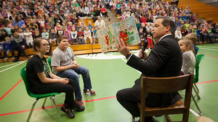 Photo of a man in a suit reading to an audience of children in a school gymnasium
