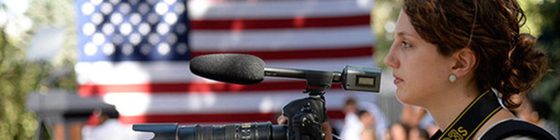 Photo of a camera woman holding a camera in front of her with an American flag in the background