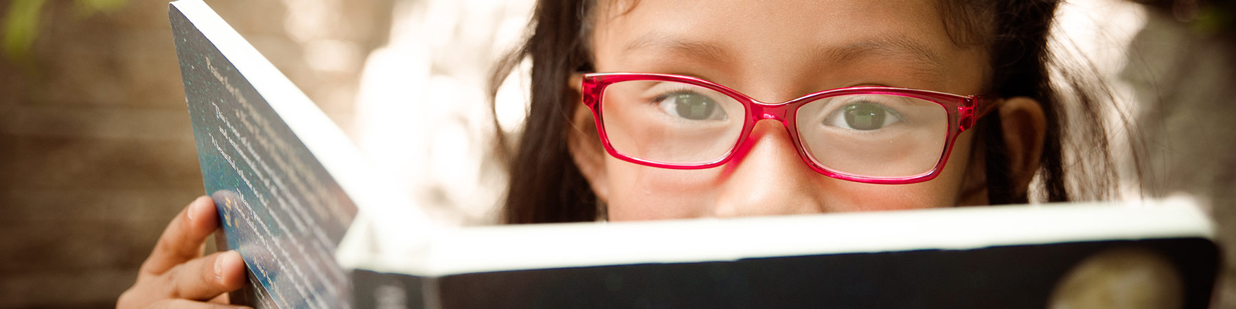 Photo of a child with red glasses peering over the top of a book