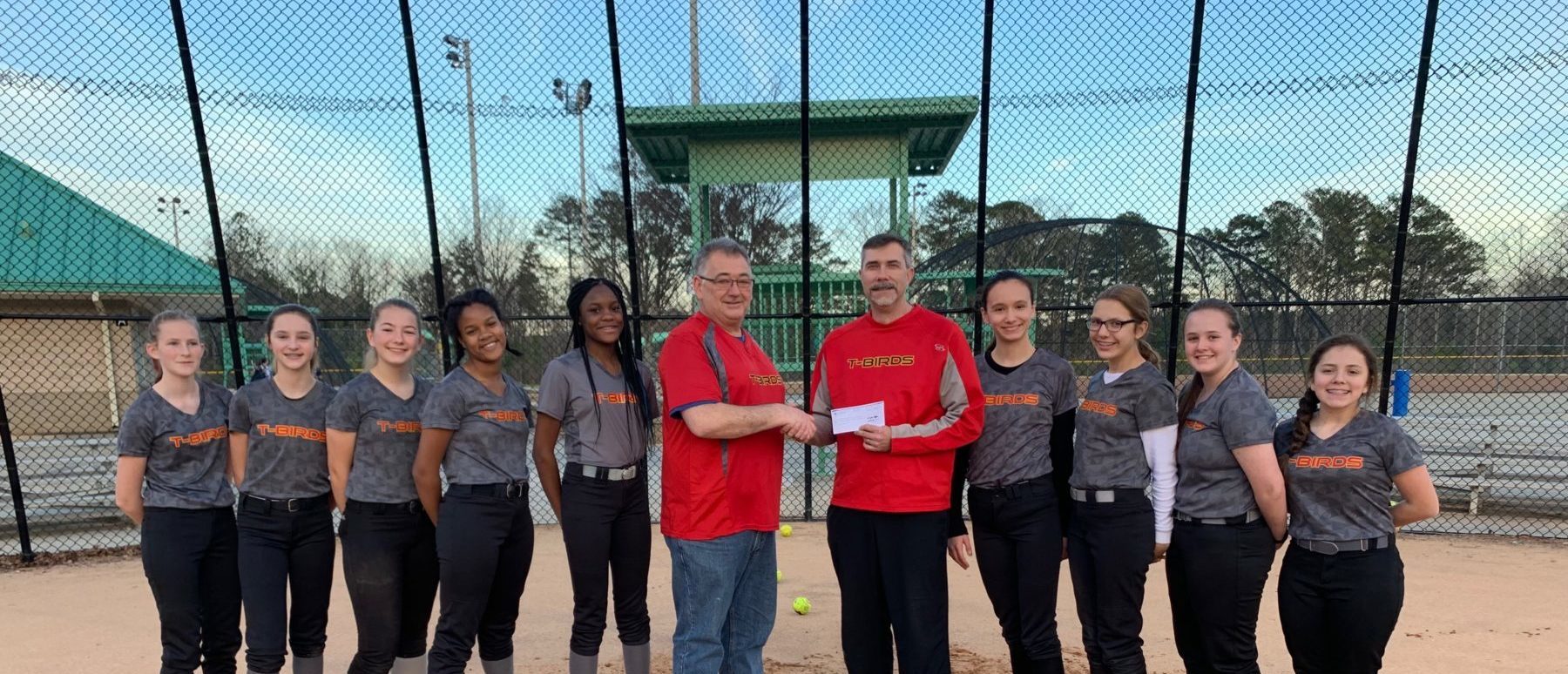 Picture of a women's softball team standing in a line with two coaches at the center shaking hands and holding a check