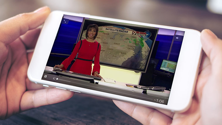 Image of two hands holding a mobile phone sideways as a female news reporter in a red dress talks about the weather on the screen