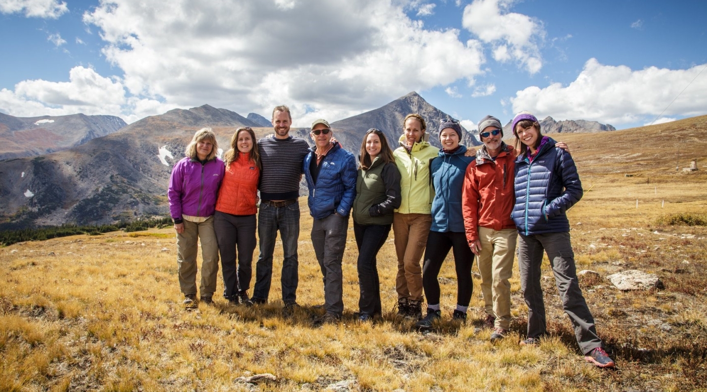 Photo of a group of men and women standing in a brownish-yellow field with huge mountains in the background