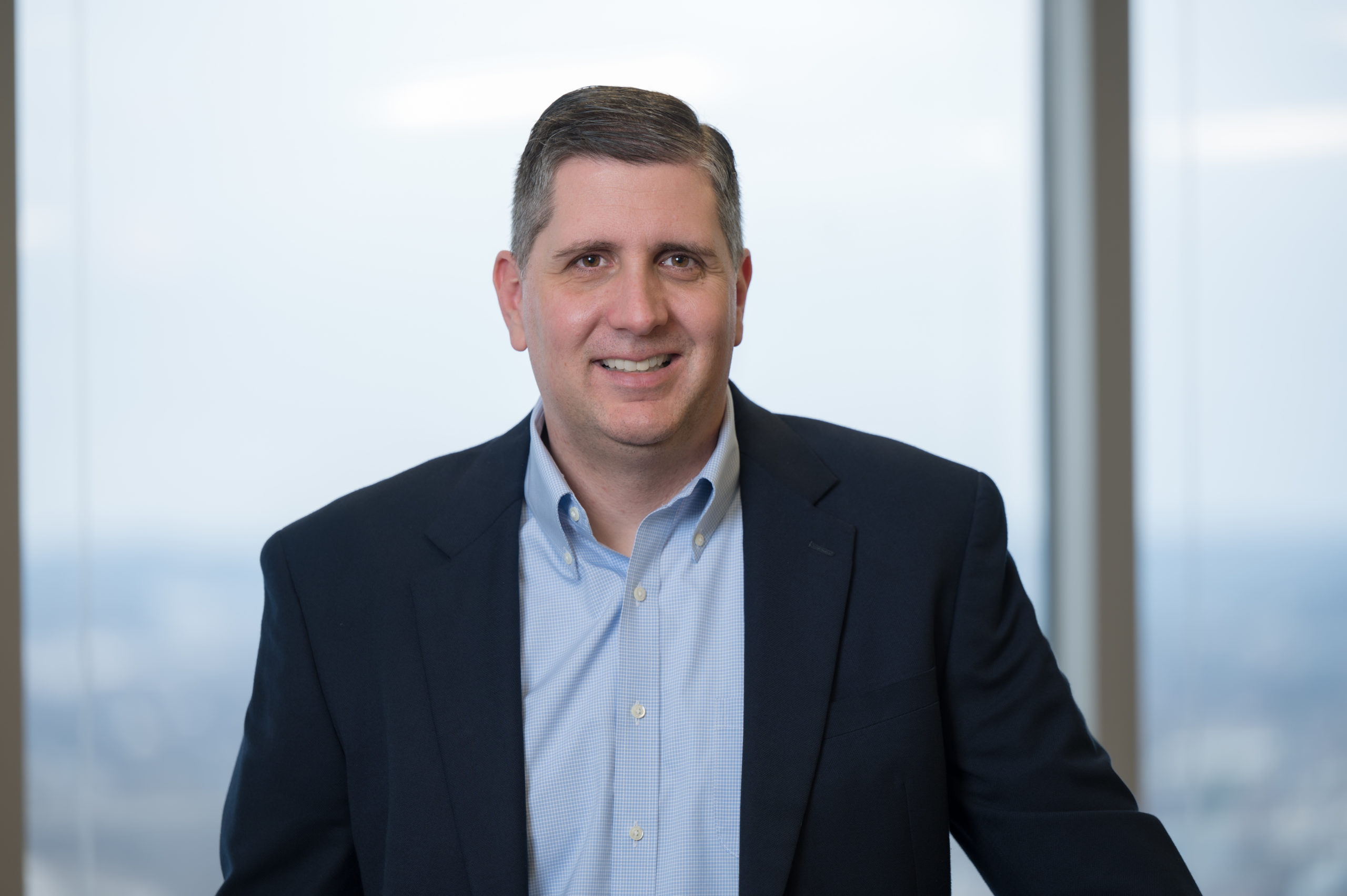 Photo of Jason Combs, chief financial officer for The E.W. Scripps Company wearing a black suit with the light blue shirt standing in front of a window
