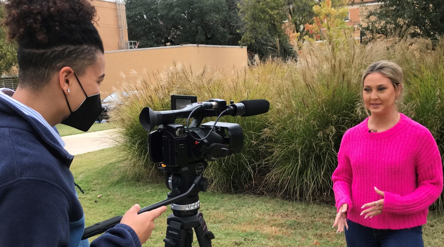 Image of a man wearing a mask over his face and videoing a female reporter in a pink sweater as she gives the news outdoors