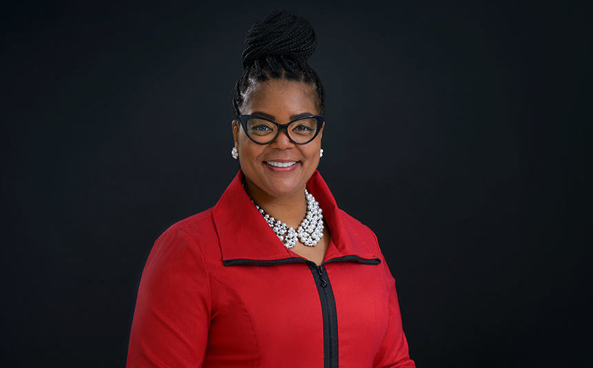Photo of Ms. Danyelle smiling and wearing a red jacket while standing in front of a black background