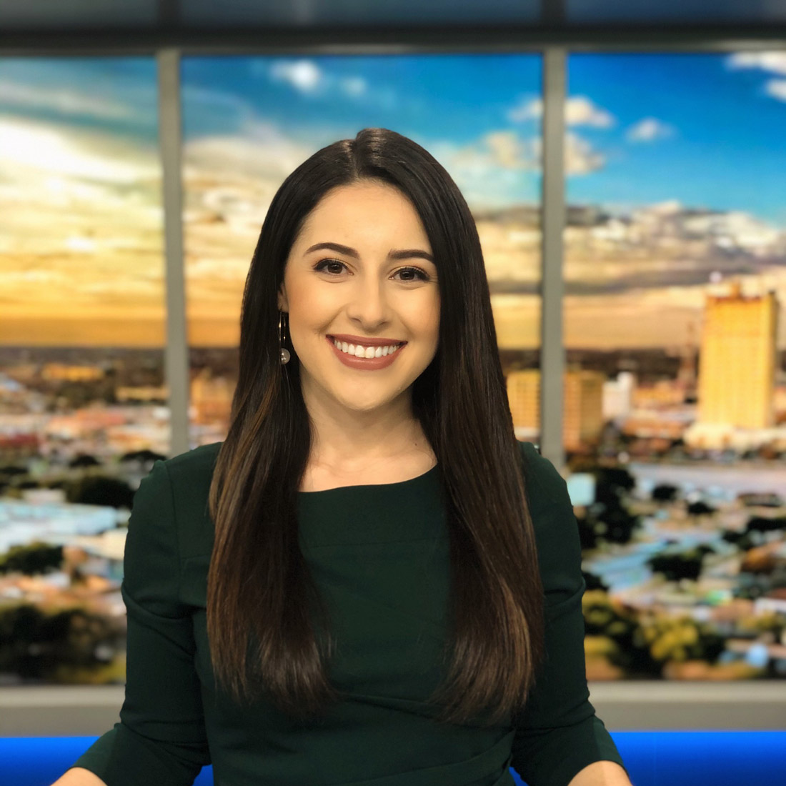 Photo of a female news reporter wearing a dark green dress and smiling in front of a city background