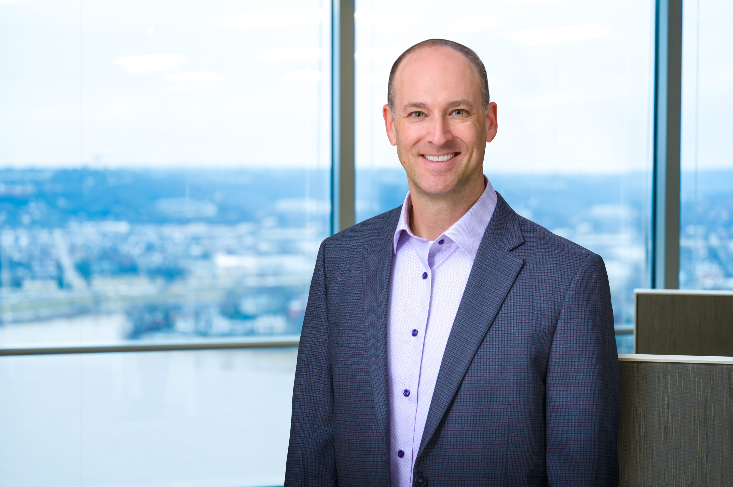 Photo of Adam Symson, president and CEO of The E.W. Scripps Company wearing a blue suit jacket with a light purple shirt standing in front of a window