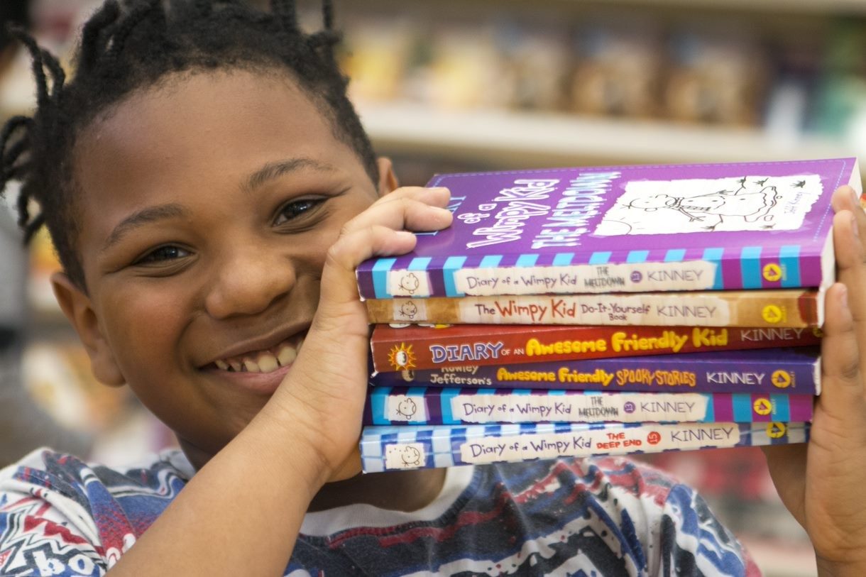 Photo of a young boy smiling and holding a stack of Diary of a Wimpy Kid series books on his shoulder
