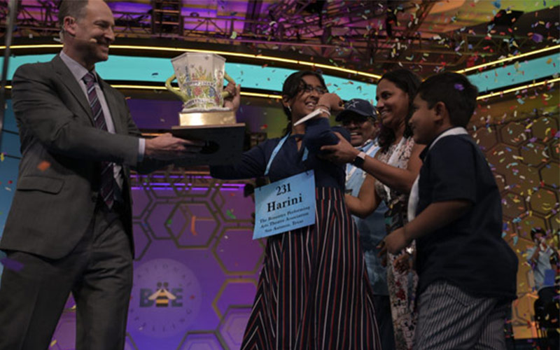A photo of a woman crying and accepting a trophy from a man after winning a spelling bee with confetti flying around the room and her family celebrating her