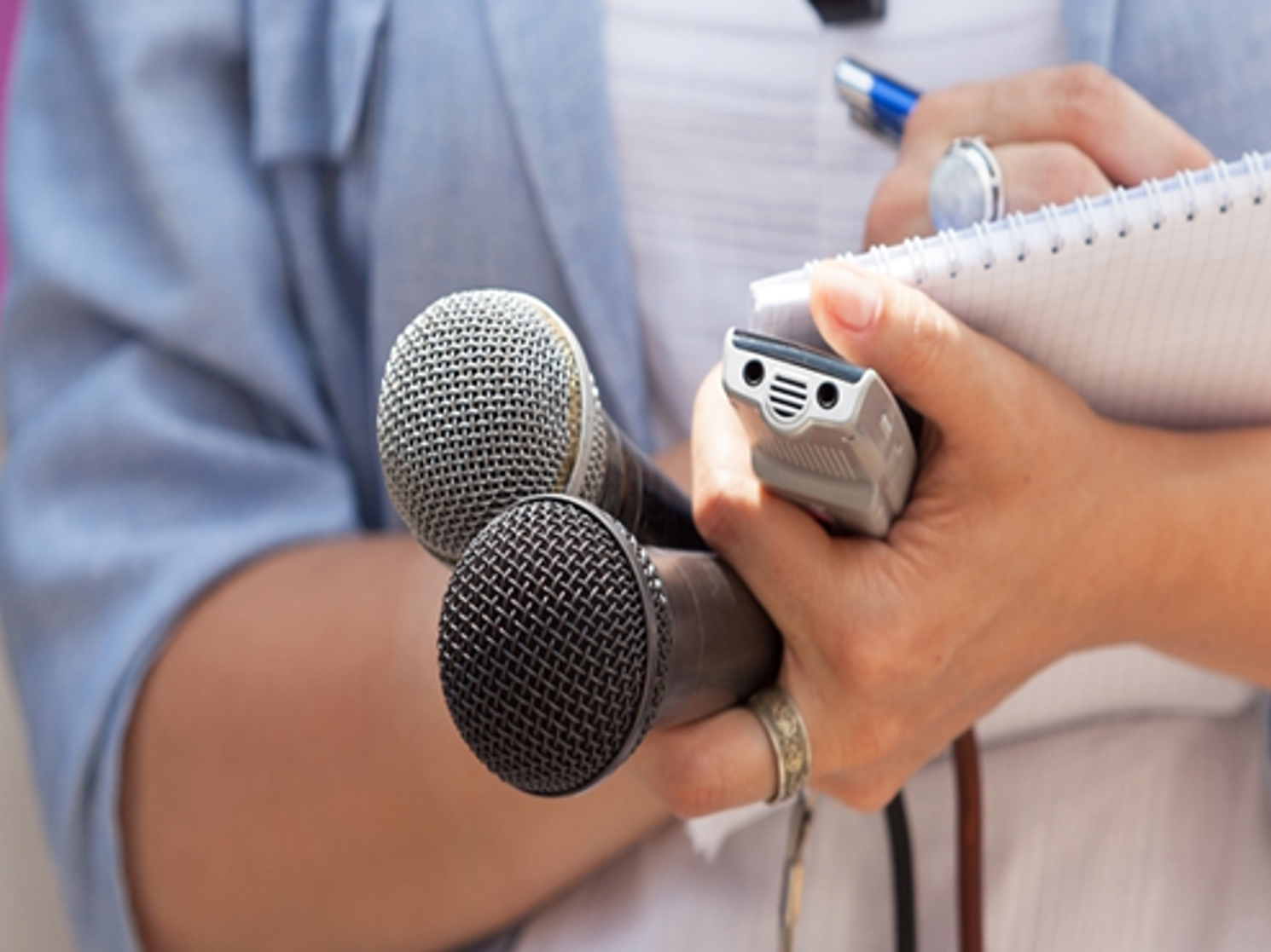 Photo of a reporter writing in a notepad with one hand and using their other hand to hold two different microphones and a recording device
