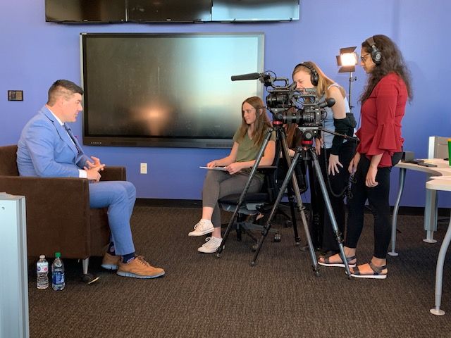 Photo of a man in a blue suit being interviewed, recorded, and listened to by three women