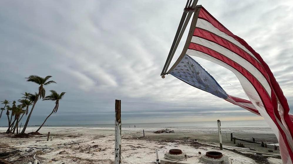 Photo of a beach after a hurricane with an American flag torn and hanging down off of a house's roof