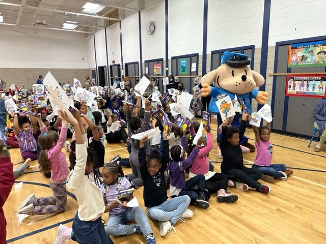 Photo of small children sitting on a gymnasium floor while they hold up colored-in pages of a coloring sheet with a police dog mascot on it while the mascot stands beside them