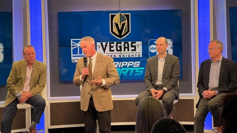 Photo of four men in tan, grey, and black dress suits sitting on a stage as one talks into a microphone with a Vegas Golden Knights graphic behind him on the Scripps Sports broadcasting screen