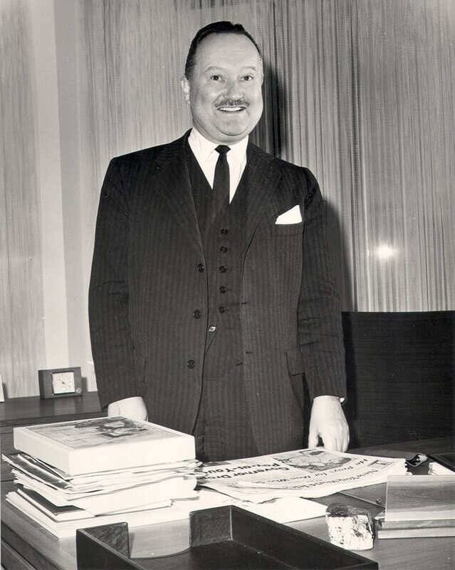 Black and white photo of a man in a striped suit smiling by his desk covered with newspapers