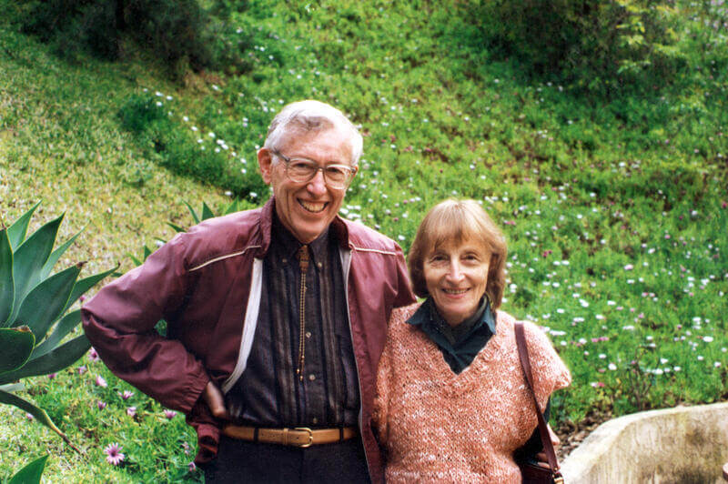 Photo of a man and woman standing by a hillside covered with green grass and wildflowers