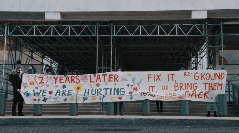 Photo of people holding up a hand-painted sign that says "2 YEARS LATER WE ARE HURTING. FIX IT, RE-GROUND IT, OR BRING THEM BACK. BOEING 737 MAX 34C LOVED ONES."
