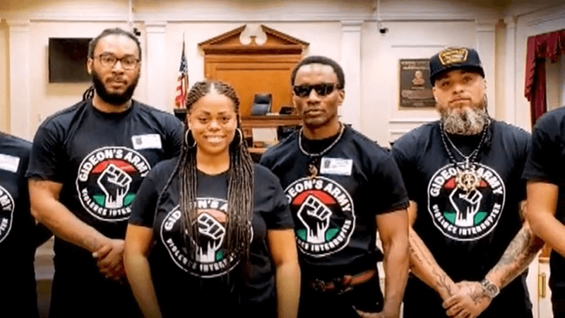 Photo of three men and a woman wearing black shirts with red, green, and white images of a fist rising up with font that says "Gideon's Army"