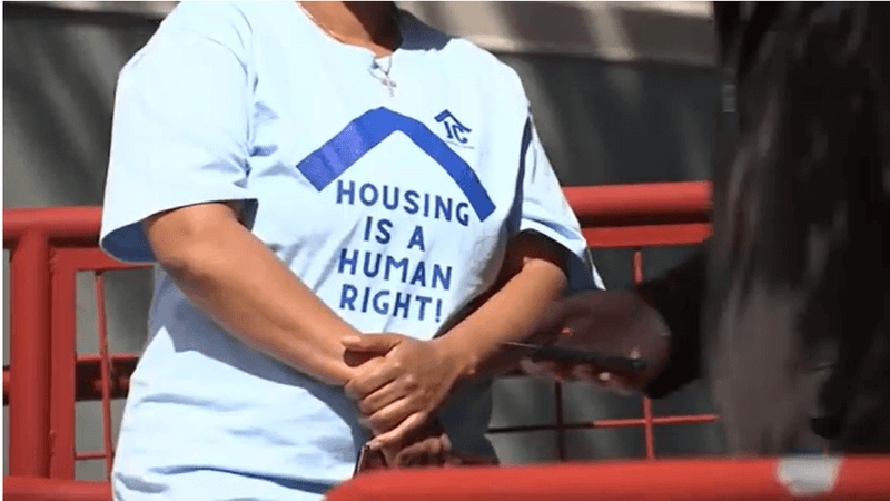 Close up image of a woman's shirt that is white with blue font that says "HOUSING IS A HUMAN RIGHT!"